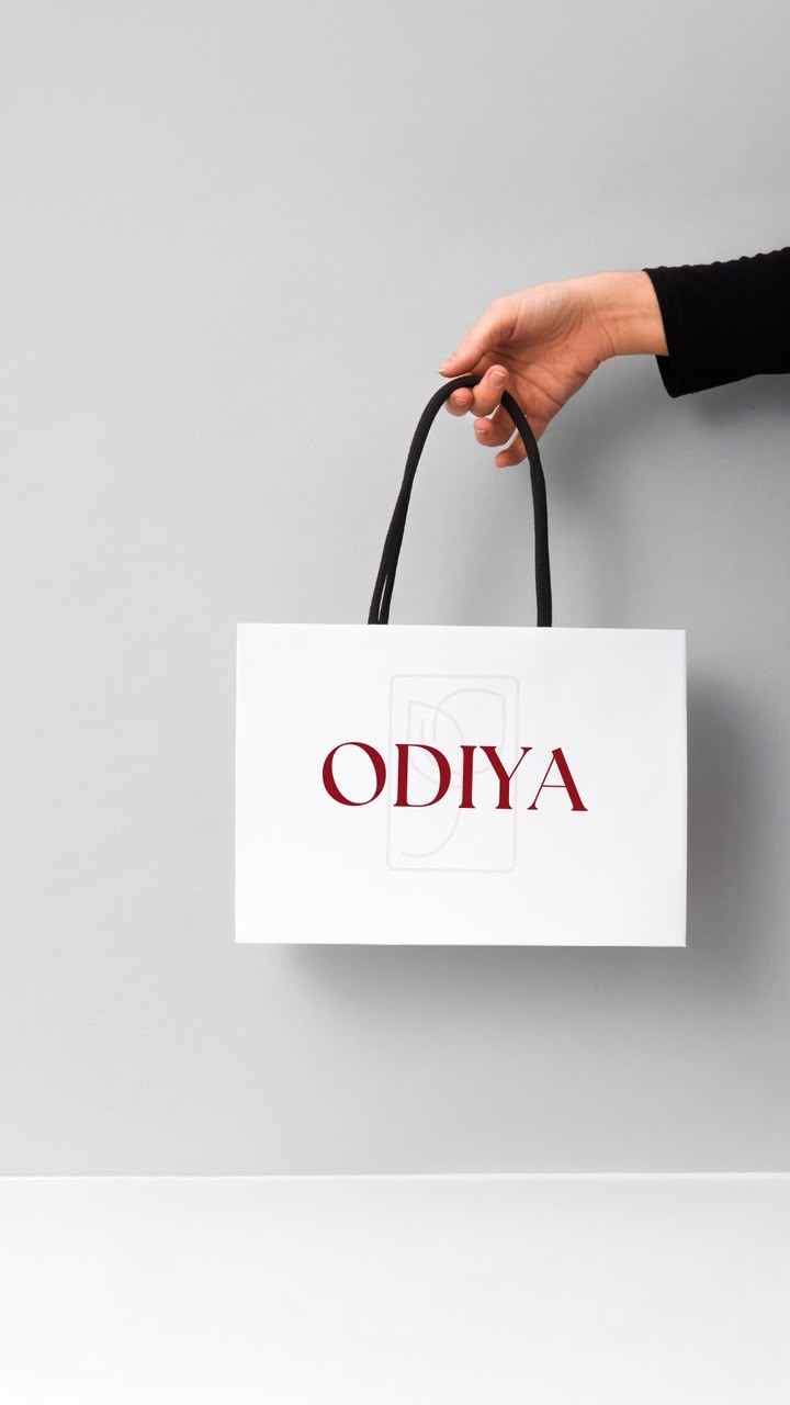 A hand holding a white ODIYA shopping bag with the oxblood wordmark, against a sage grey background.